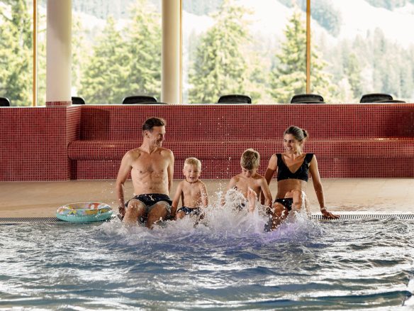 Familie mit zwei Kindern sitzt am Rand des Panoramahallenbads und lässt das Wasser spritzen, dahinter eine rote Wärmebank.