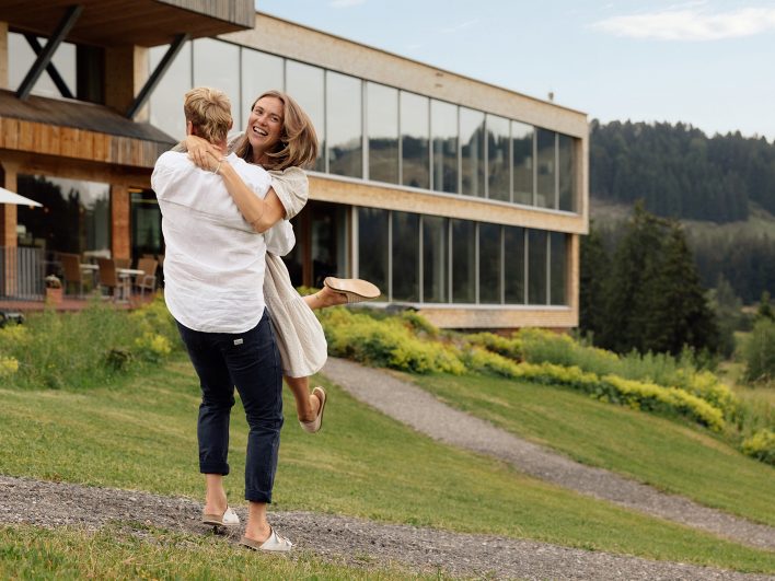 Mann hebt eine lachende Frau vor dem Almhotel Hochhäderich hoch und dreht sich mit ihr auf dem Weg an der Wiese.