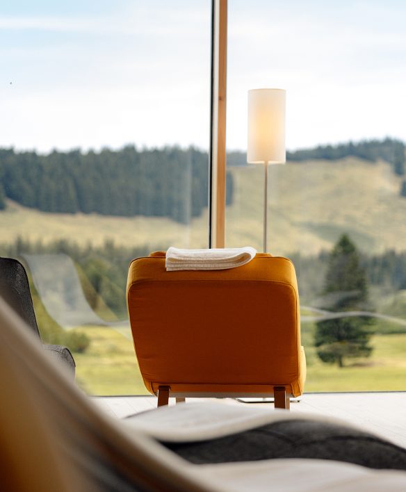 Orangefarbene Relaxliege mit aufgelegter Decke am Panoramafenster des Ruheraums, Blick auf Almwiese und Wald.