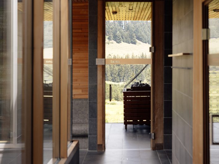 Blick durch die Glastüren im AlmSpa in die Saunen, dahinter der Saunaofen und die Fensterfront zur Almwiese.