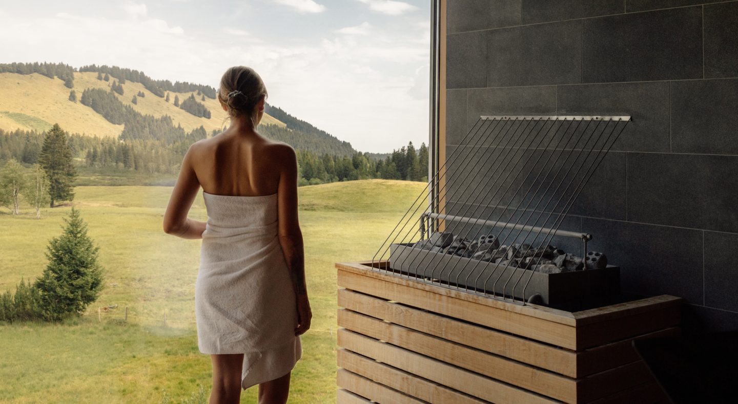 Frau im Saunatuch steht am Panoramafenster der Sauna und schaut auf die Almwiesen, rechts der Saunaofen mit heißen Steinen.