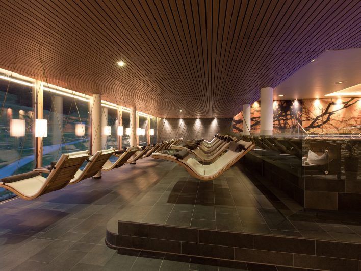 Ruheraum im Adults-only AlmSpa des Almhotels Hochhäderich mit einer Reihe frei hängender Schwebeliegen aus Holz, gedämpftem Licht und großen Fenstern zur Almlandschaft.