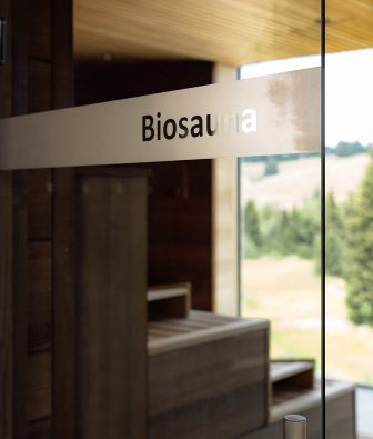 Glastür mit der Aufschrift Biosauna, dahinter dunkle Holzbänke und ein Fenster mit Blick ins Grüne.