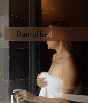 Frau im Handtuch öffnet die beschlagene Glastür mit der Aufschrift Dampfbad von innen.