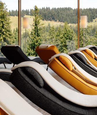 Geschwungene Relaxliegen in Grau und Gelb stehen im Ruheraum vor der Fensterfront zum Wald.