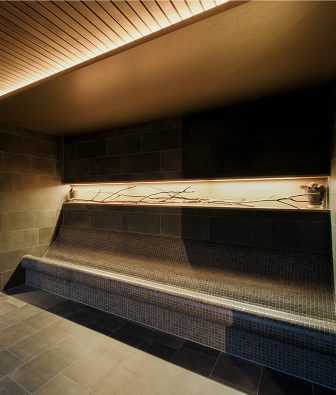 Geschwungene, mit dunklem Mosaik geflieste Wärmebank im AlmSpa, darüber eine warm beleuchtete Nische mit Ästen als natürlicher Dekoration.