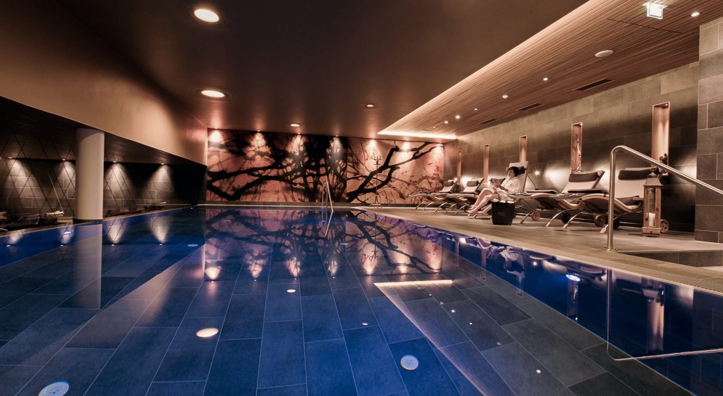 Nacktschwimmbecken im Adults-only AlmSpa mit dunkel schimmerndem Wasser, großem Wandbild aus Baumästen und einer Frau im Bademantel, die auf einer Holzliege liest.