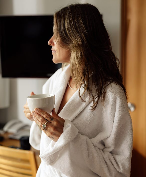Frau im weißen Bademantel steht mit einer Tasse in der Hand im Hotelzimmer und schaut nach draußen.