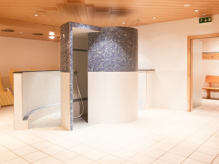 Runde Erlebnisdusche mit blauem Glasmosaik im FamSpa des Almhotels Hochhäderich, daneben ein gefliester Kneipp-Gang und gestreifte Liegen im Ruhebereich.