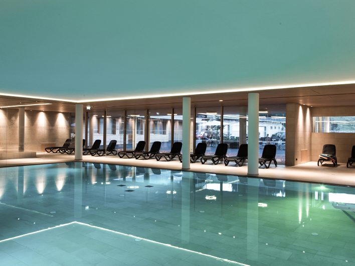 Türkis schimmerndes Schwimmbecken im Panorama-Hallenbad, gesäumt von schwarzen Liegen und hellen Säulen, durch die Glasfront fällt der Blick auf den Innenhof.