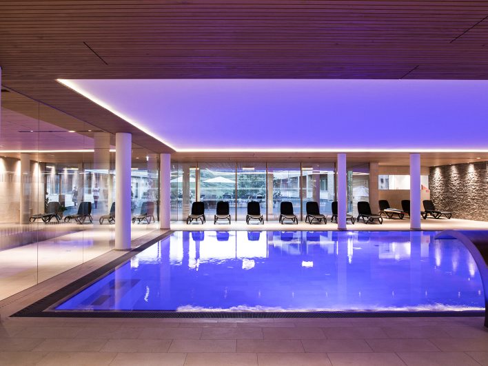 Panorama-Hallenbad im Almhotel Hochhäderich in violettem Licht mit Holzdecke, Natursteinwand und einem Wasserspeier am Beckenrand, der sich zur Nackenmassage eignet.