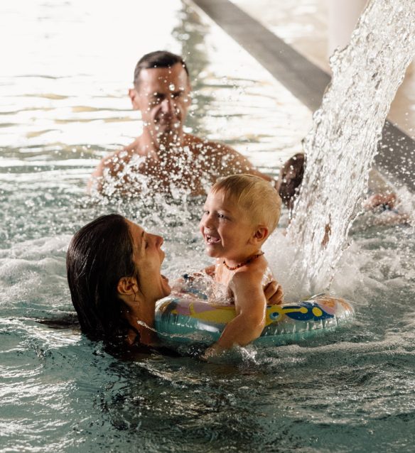 Mutter hält ihr lachendes Kleinkind mit Schwimmreifen unter dem Wasserfall im Hallenbad, dahinter schwimmt der Vater.