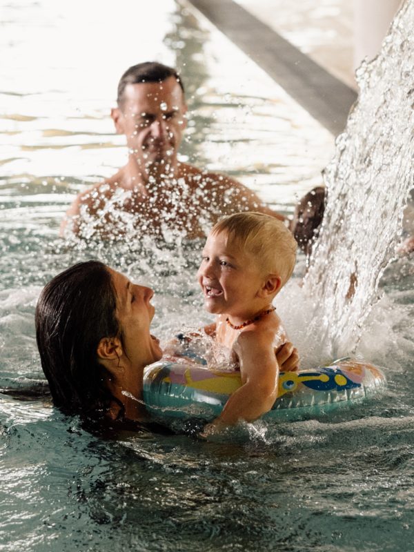 Mutter hält ihr lachendes Kleinkind mit Schwimmreifen unter dem Wasserfall im Hallenbad, dahinter schwimmt der Vater.