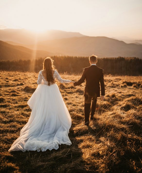 Brautpaar geht Hand in Hand über eine Bergwiese im goldenen Abendlicht, die Braut im weißen Spitzenkleid mit langer Schleppe, vor ihnen eine weite Hügellandschaft.