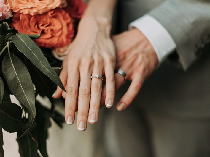 Hände eines frisch verheirateten Paares mit Eheringen übereinandergelegt, daneben ein Brautstrauß mit orangen Rosen.