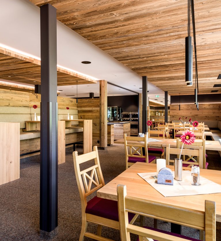 Gastraum des Johann & Frieda am Almhotel Hochhäderich mit Altholzdecke, gemütlichen Sitznischen und Holzstühlen mit roten Sitzpolstern, auf jedem Tisch eine rote Blume.