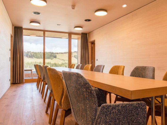 Kleiner Seminarraum mit langem Holztisch, gepolsterten Stühlen in Ocker und Graublau und raumhohem Fenster in die Berglandschaft.