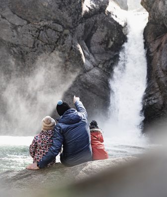 Erwachsener mit zwei Kindern sitzt auf einem Felsen vor den Buchenegger Wasserfällen im Allgäu und zeigt auf das herabstürzende Wasser