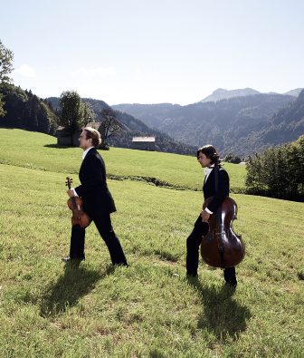 Zwei Musiker in dunklen Anzügen mit Geige und Cello gehen über eine Bergwiese bei Schwarzenberg, Sinnbild für das Musikfestival Schubertiade
