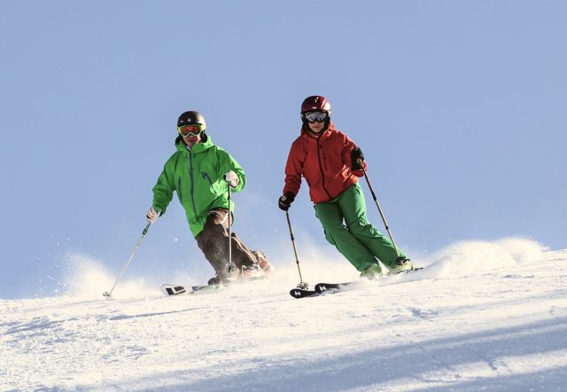 Zwei Skifahrer in grüner und roter Jacke carven nebeneinander durch den Pulverschnee, der bei jedem Schwung aufstiebt.