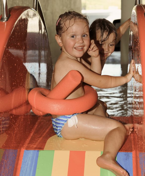 Kleines Mädchen mit Schwimmnudel sitzt strahlend oben auf der Regenbogenrutsche im Kinderbecken, hinter ihm wartet schon das nächste Kind.