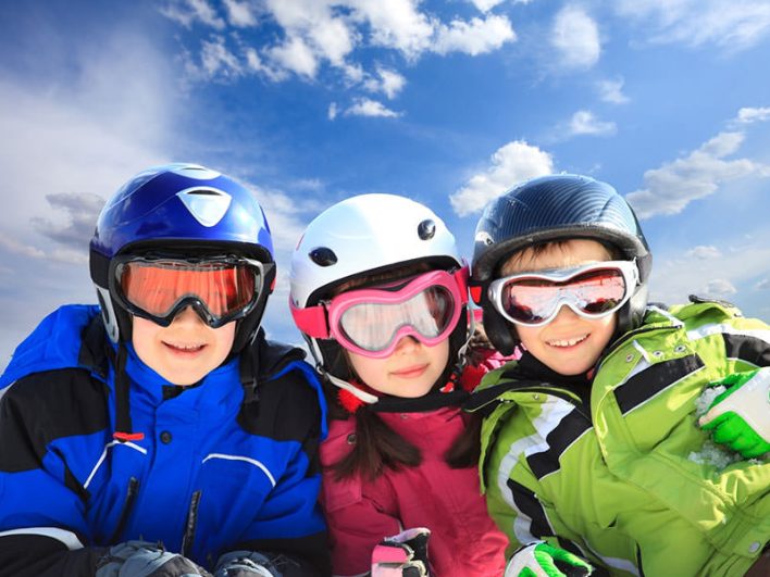 Drei Kinder in bunten Winterjacken mit Skihelmen und Skibrillen.